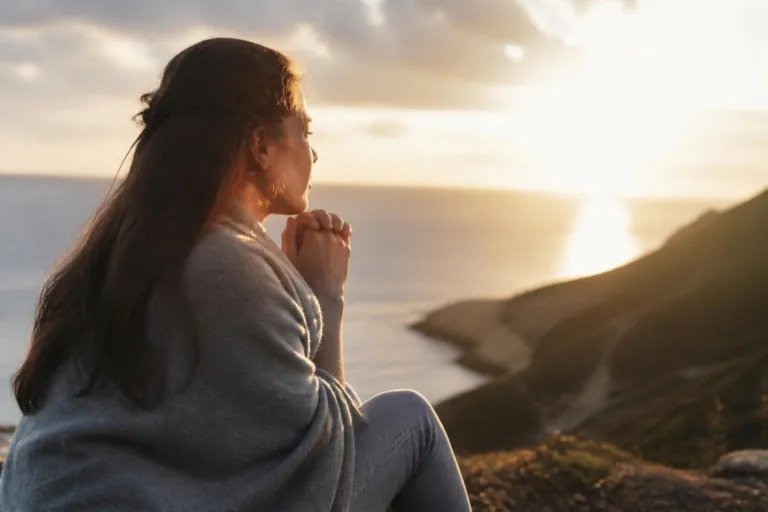 Woman sitting on a rocky hillside at sunset, hands clasped in prayer, gazing toward the sea.