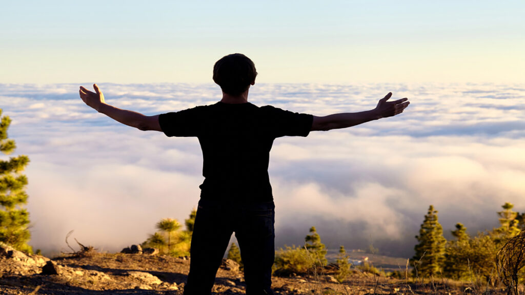 Person stands on rocky hill with arms outstretched, facing a sea of clouds at sunrise or sunset over a forested valley