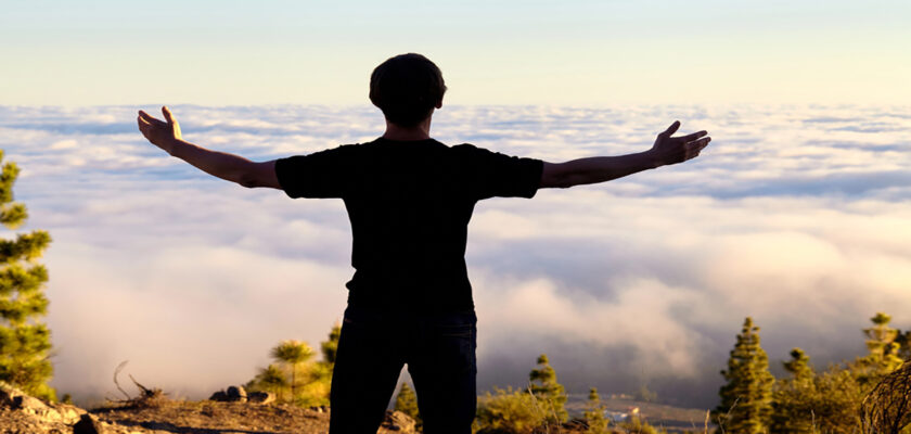 Person stands on rocky hill with arms outstretched, facing a sea of clouds at sunrise or sunset over a forested valley