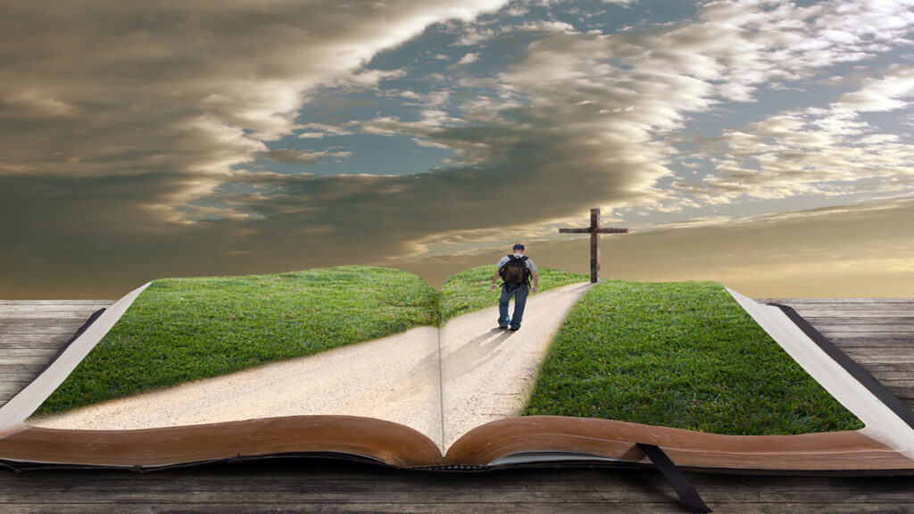 Open book on a wooden surface with a grassy path unfolding from its pages toward a distant cross as a man walks along.