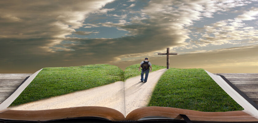 Open book on a wooden surface with a grassy path unfolding from its pages toward a distant cross as a man walks along.
