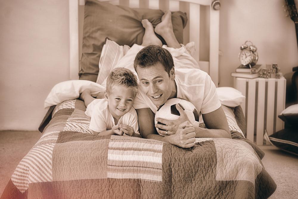 Father and young son lying on a bed, smiling and holding a soccer ball between them.