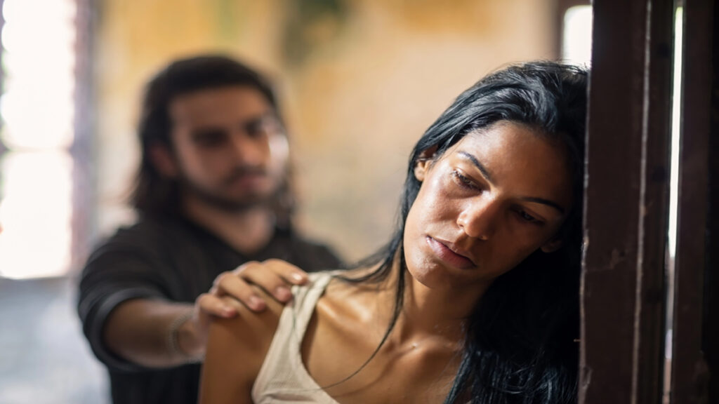Woman leaning against a wooden frame with a somber expression while a man behind her places a hand on her shoulder.