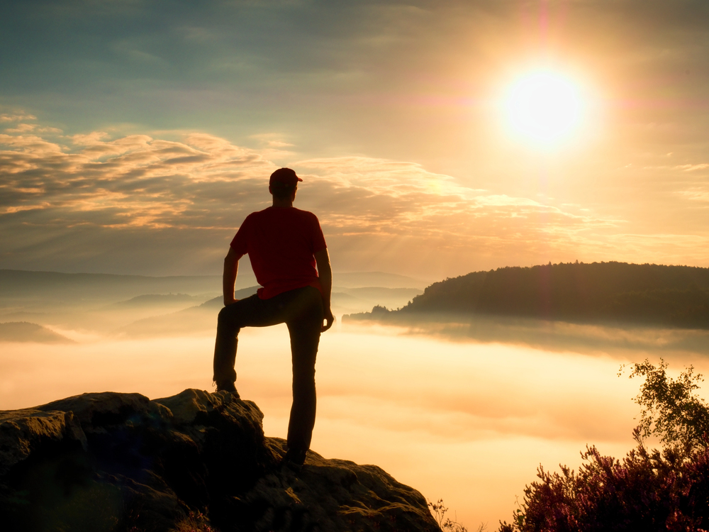 Depositphotos_120236932_S - Renuevo De Plenitud Person standing on a rocky cliff at sunrise, overlooking a misty valley and distant hills.