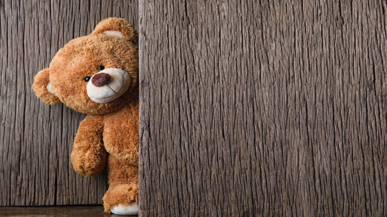 Brown teddy bear peeking from behind a dark wooden plank wall or door, with only half its body visible and a friendly face.