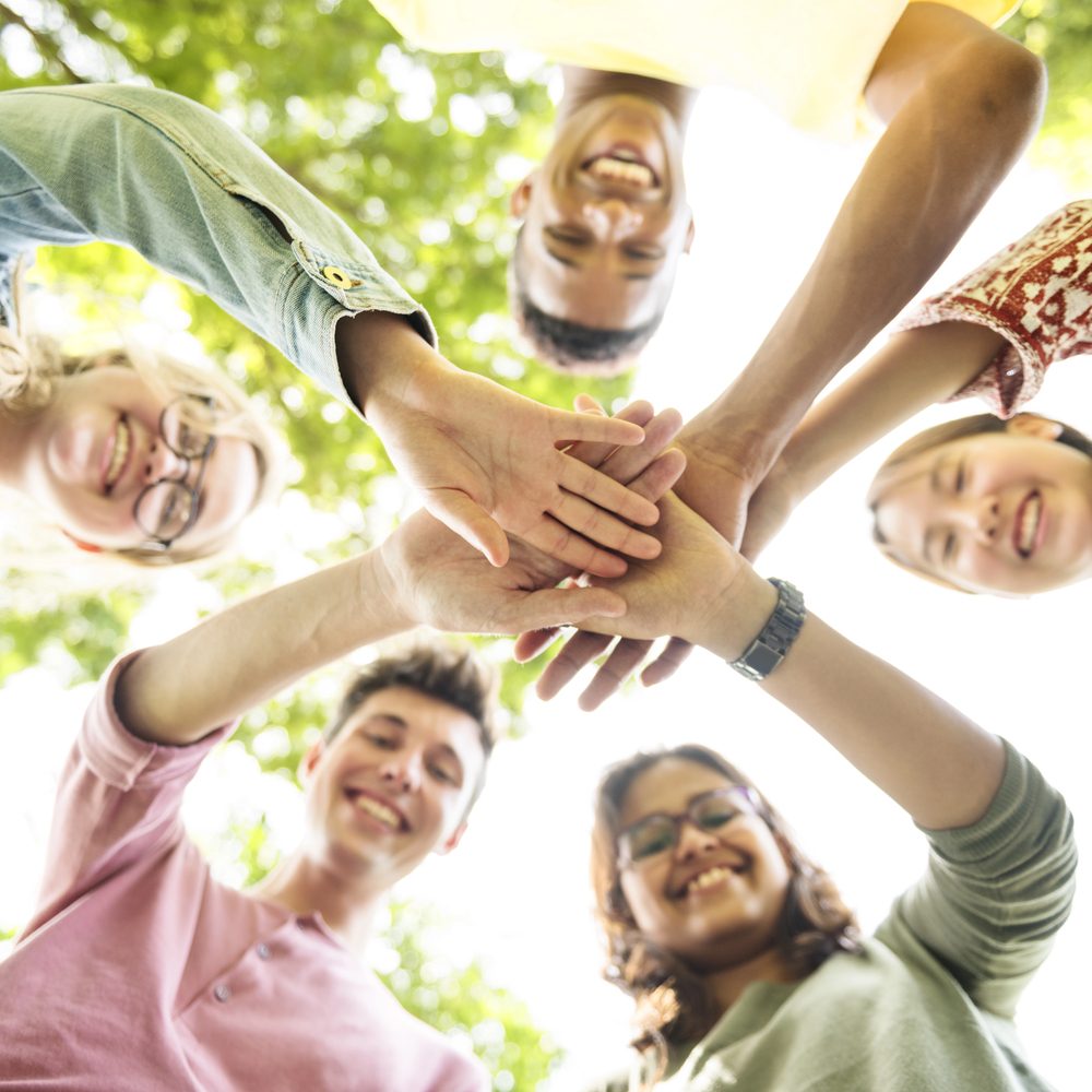 Group of diverse young adults standing in a circle and stacking hands in the center outdoors, showing teamwork and unity.