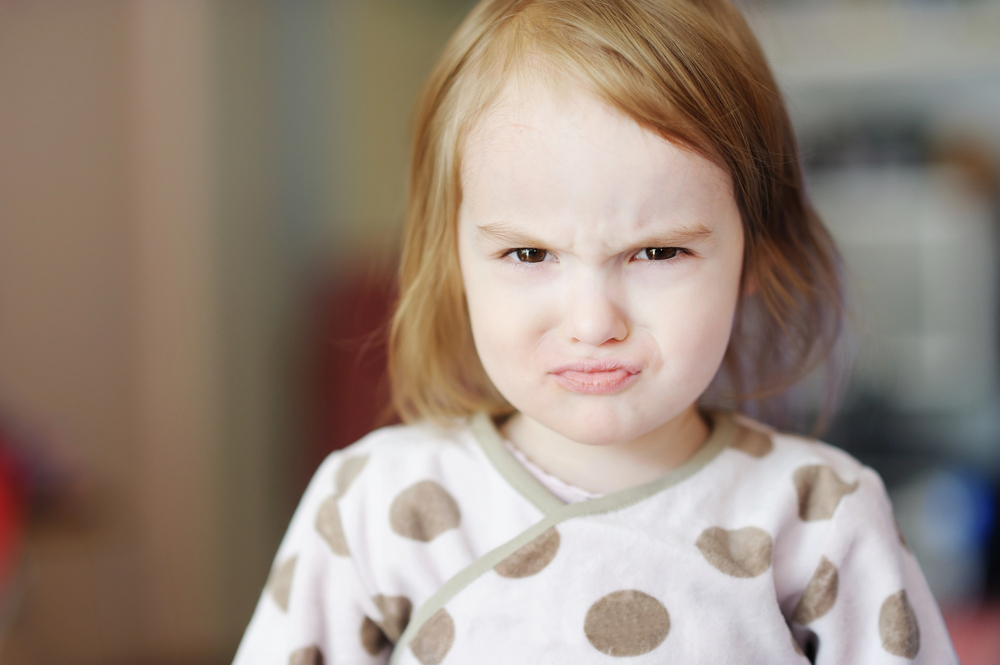 Young child with blond hair making a pouty face, wearing a white polka-dot top with brown circles.