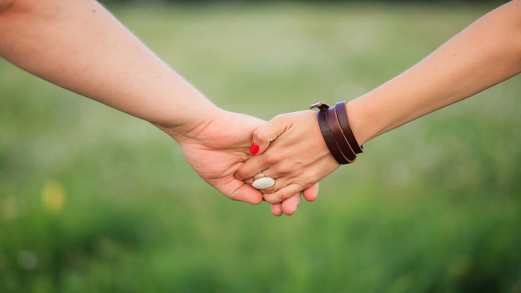 Two people hold hands in a supportive handshake outdoors with a green field in the background in soft focus.