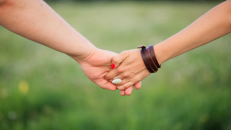 Couple Holding Hands - Renuevo De Plenitud Two people hold hands in a supportive handshake outdoors with a green field in the background in soft focus.