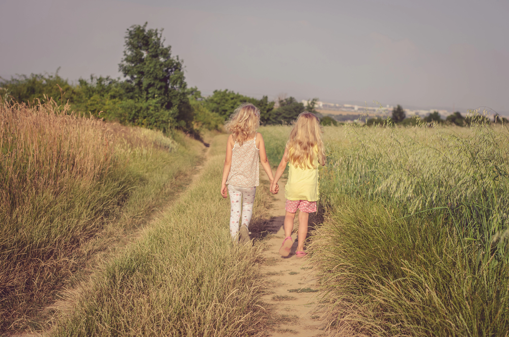 Two young girls walk hand-in-hand along a dirt path between tall grasses on a sunny day.