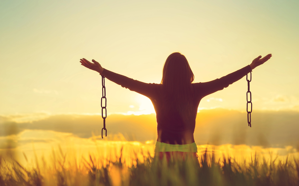 Depositphotos_151871670_S - Renuevo De Plenitud Silhouette of a person with arms wide open in a sunlit field, chains hanging from both wrists, symbolizing freedom at sunset.