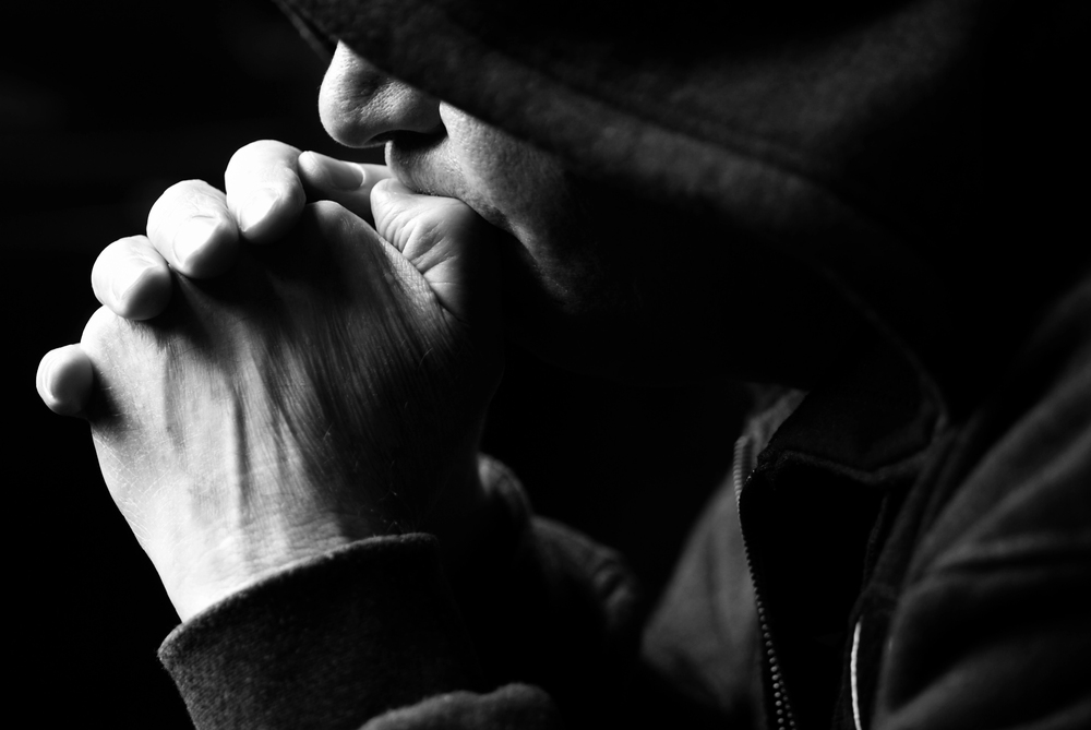 Close-up of a person in a hooded sweatshirt with hands pressed together in prayer, in black and white.