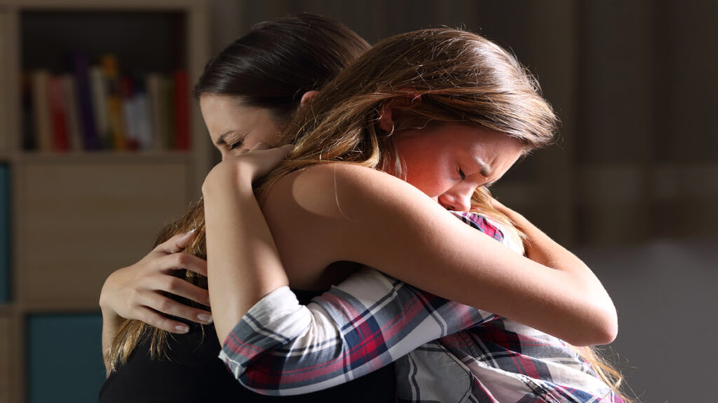 Two friends share a tight hug; one woman appears distressed while the other consoles her indoors