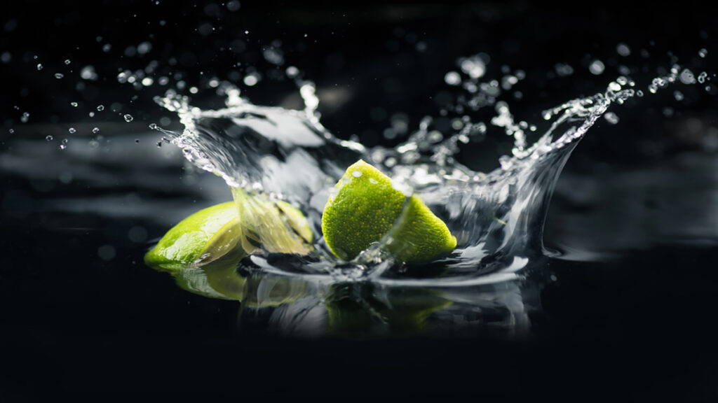 Lime wedges splashing into dark water with droplets arcing in midair against a black background.