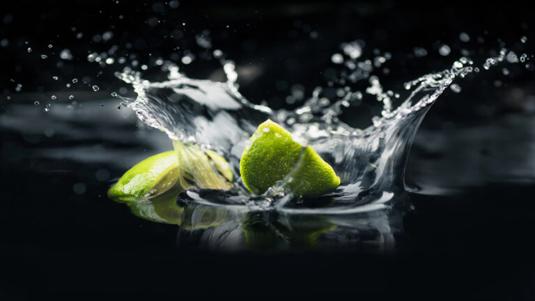 Lime wedges splashing into dark water with droplets arcing in midair against a black background.