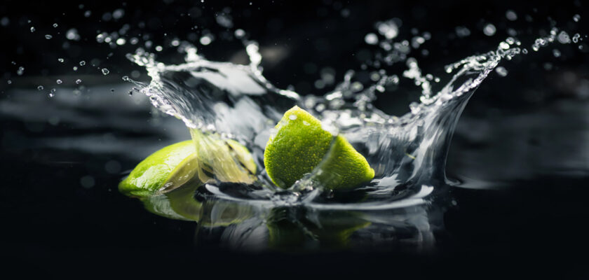 Lime wedges splashing into dark water with droplets arcing in midair against a black background.
