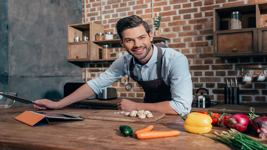Smiling man in a blue shirt and apron leans on a wooden kitchen counter with a knife and fresh vegetables nearby.