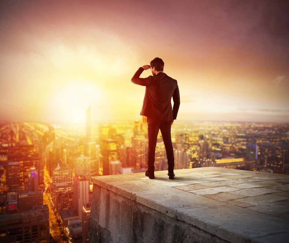 Man in a suit stands on a rooftop ledge, overlooking a sunlit city skyline at sunset, surveying the horizon.