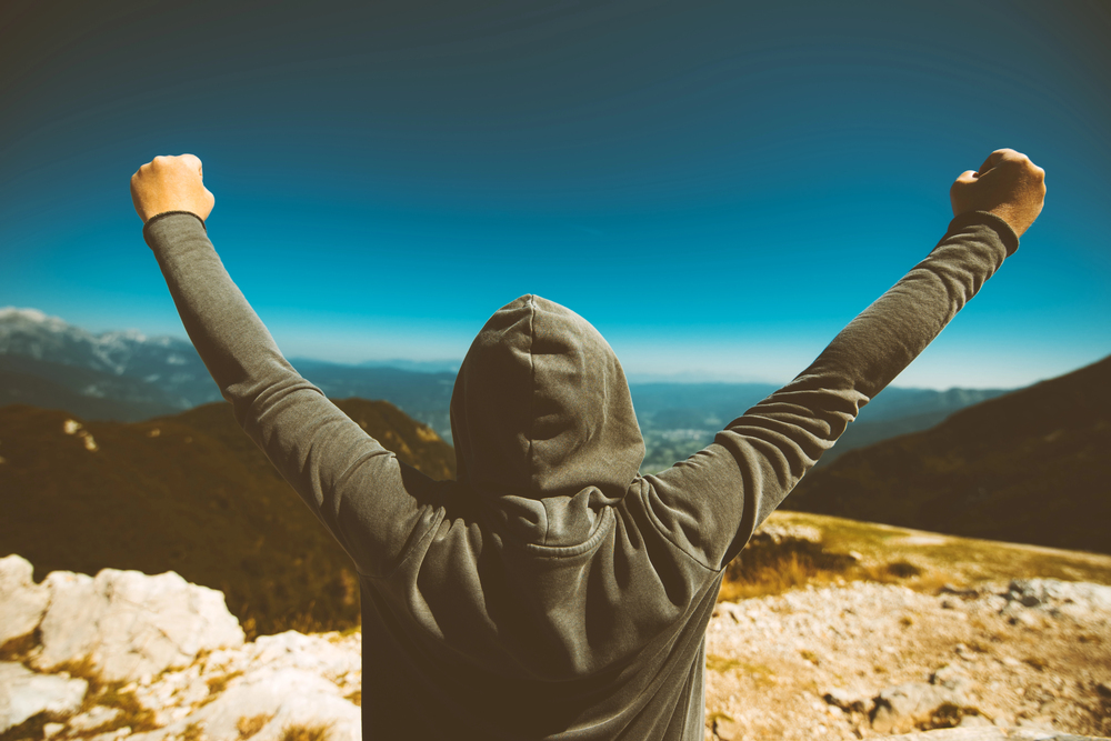 Back view of a person in a gray hoodie with arms raised in triumph at a rocky mountain summit under a blue sky