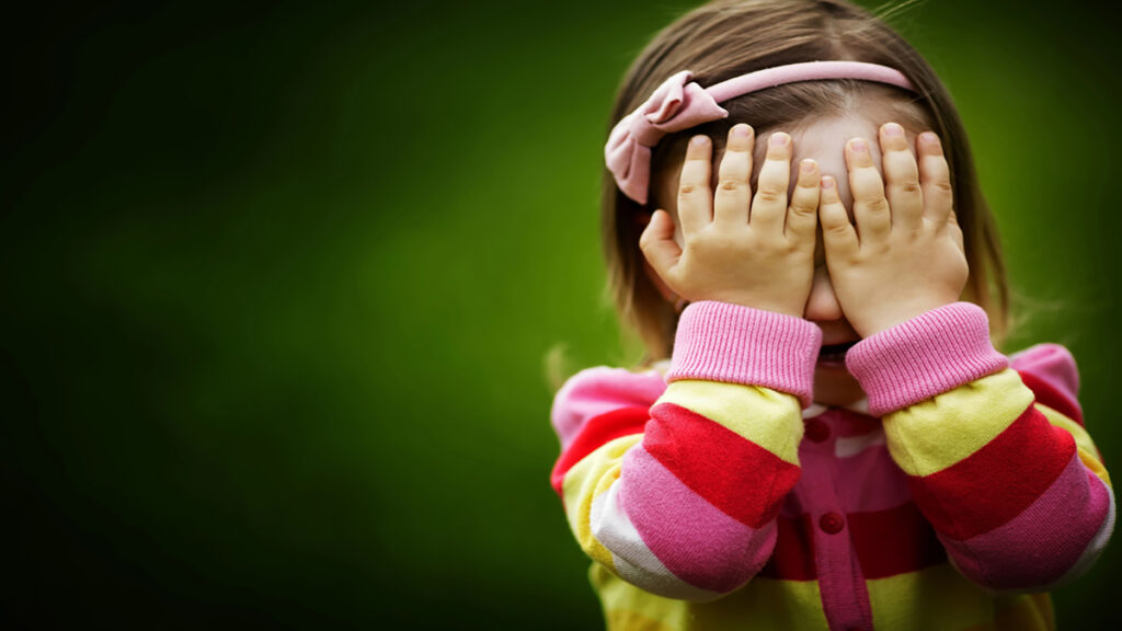 Young girl in a pink headband covers her face with both hands while wearing a colorful striped sweater outdoors, a shy/playful moment.