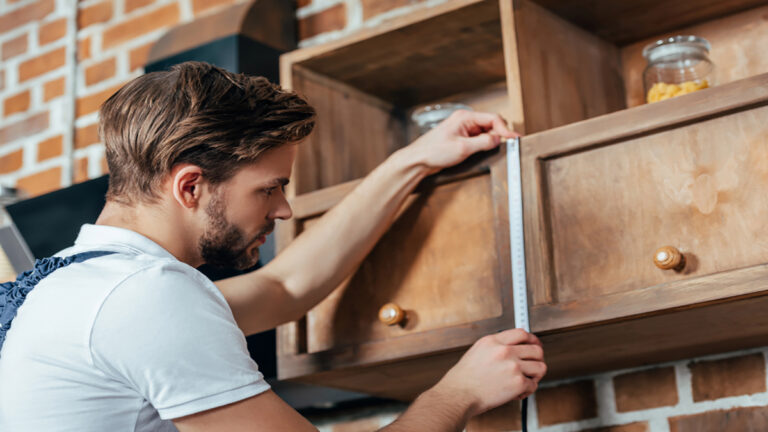 Man using a tape measure to check the height of a wooden cabinet in a workshop.