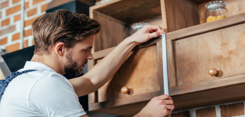 Man using a tape measure to check the height of a wooden cabinet in a workshop.