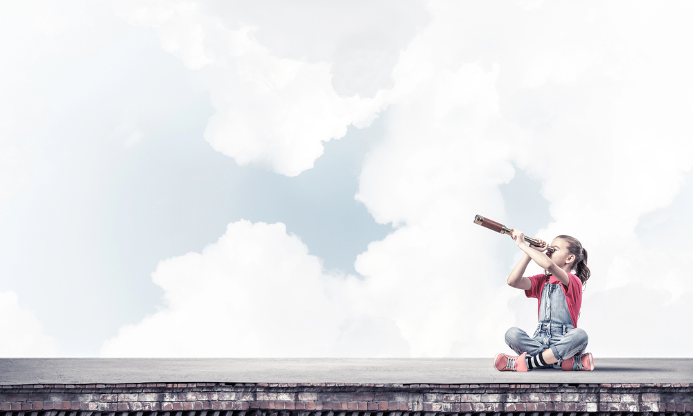 Young girl in overalls sits cross-legged on a rooftop, looking through a telescope toward the sky with clouds in the background.