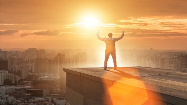 Person standing on a rooftop edge with arms raised as the sun rises over the city skyline, golden glow all around.