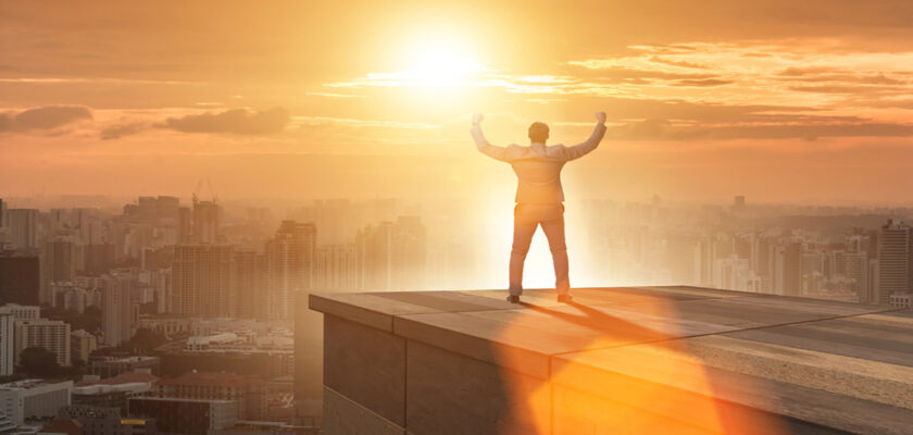 Person standing on a rooftop edge with arms raised as the sun rises over the city skyline, golden glow all around.
