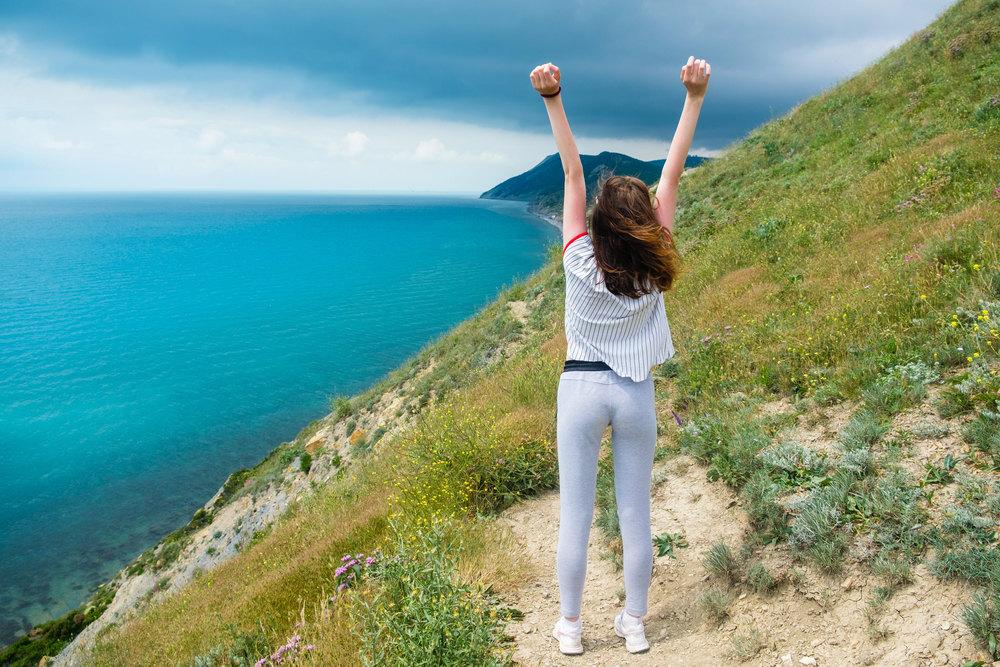 A woman on a coastal hillside raises her arms in triumph, overlooking turquoise sea and distant cliffs.