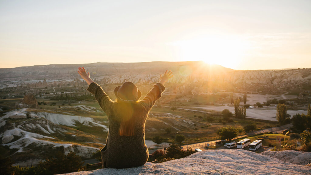 Person in a sweater and hat stands on a rocky overlook with arms raised as the sun sets over a desert-like valley.