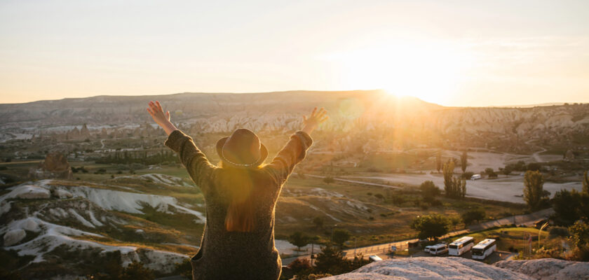 Person in a sweater and hat stands on a rocky overlook with arms raised as the sun sets over a desert-like valley.