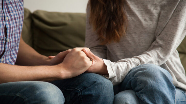 Two people sit on a couch, holding hands in a comforting embrace in a close-up.