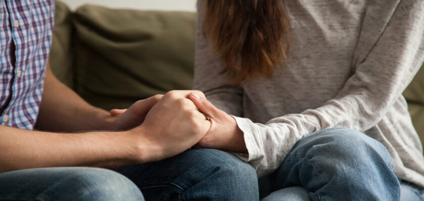 Two people sit on a couch, holding hands in a comforting embrace in a close-up.