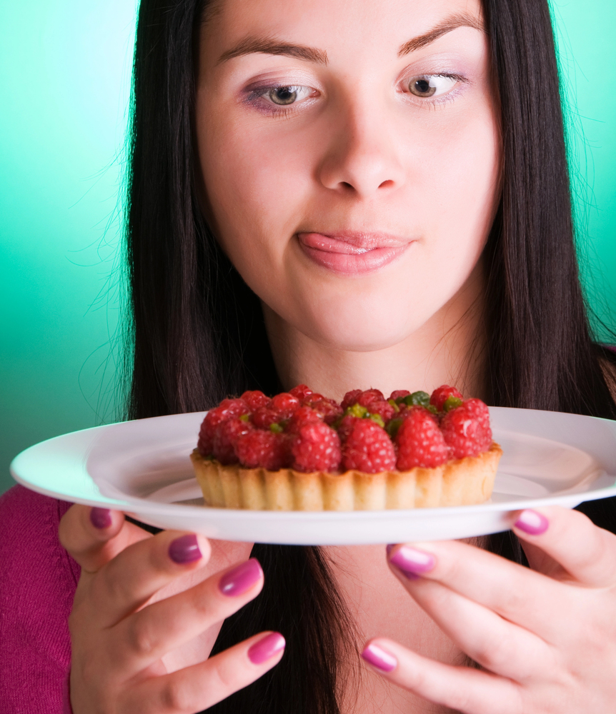 Young woman presents a raspberry tart on a white plate, looking at it with a thoughtful expression.