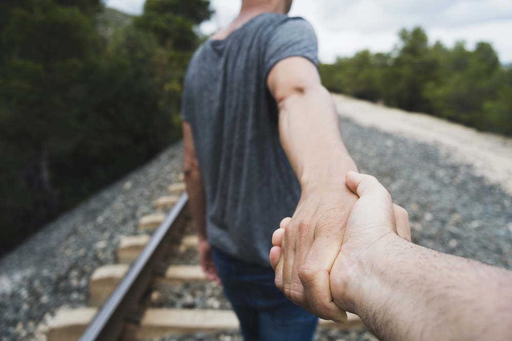 First-person view of two people holding hands while walking along a railroad track through a forested area.