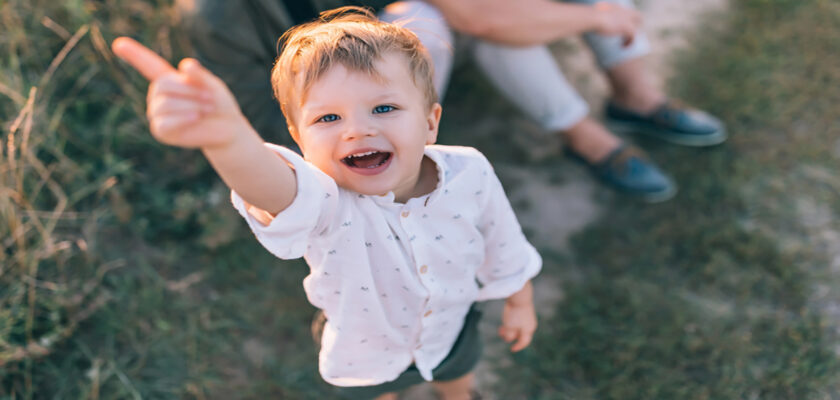 Joyful toddler points and smiles at the camera while standing on a grassy path, with an adult seated in the background.