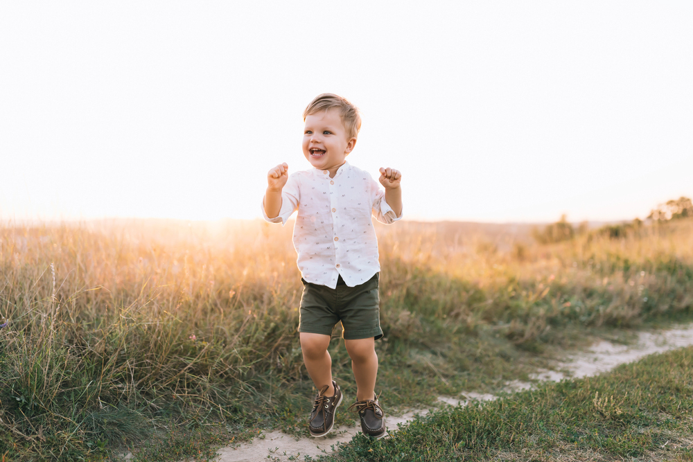 Joyful toddler running along a dirt path in a sunlit field at sunset, arms raised and a big smile on his face.