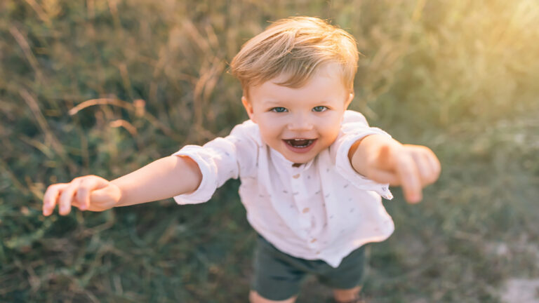 Smiling toddler reaching toward the camera with outstretched arms in a sunlit outdoor setting.