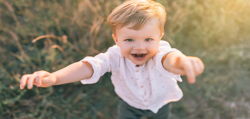 Smiling toddler reaching toward the camera with outstretched arms in a sunlit outdoor setting.