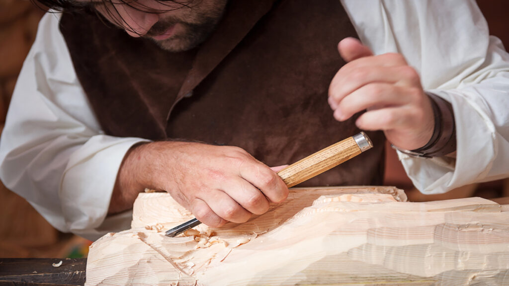 Hands of a woodworker smoothing a light block of wood with a chisel, wood shavings scattered around the work surface