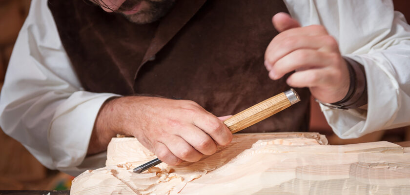 Hands of a woodworker smoothing a light block of wood with a chisel, wood shavings scattered around the work surface
