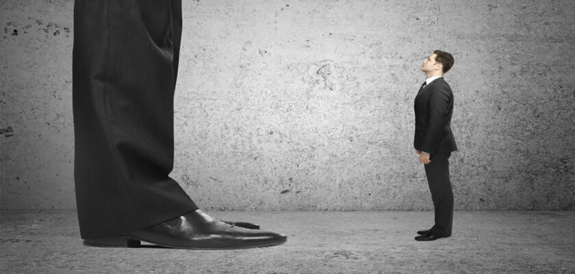 A gigantic man in a suit looms over a much smaller man in a suit, both standing on a concrete floor.