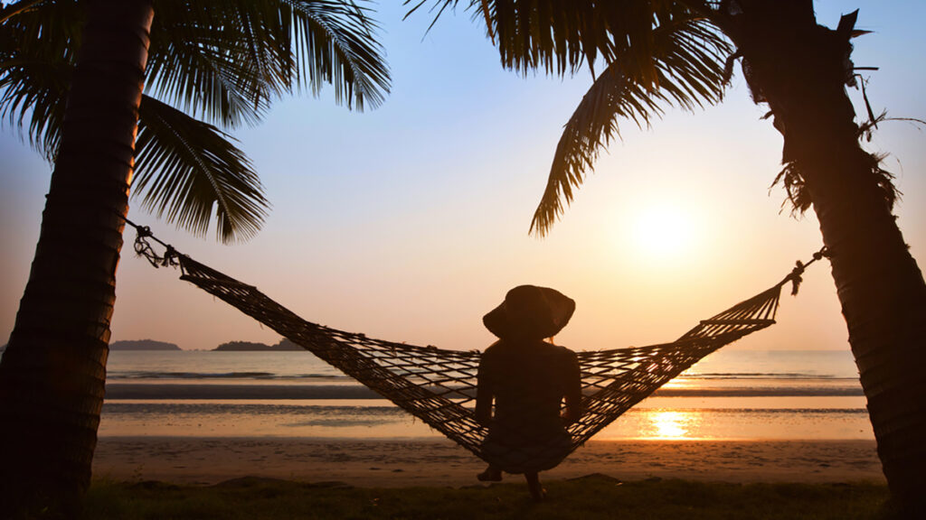 Person relaxing in a hammock strung between two palm trees on a sandy beach at sunset.