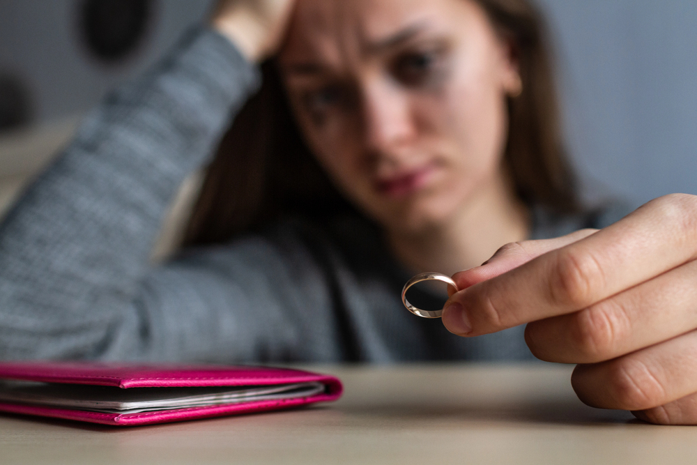 Distraught woman holding a wedding ring near the camera with a pink notebook on the table in the foreground.