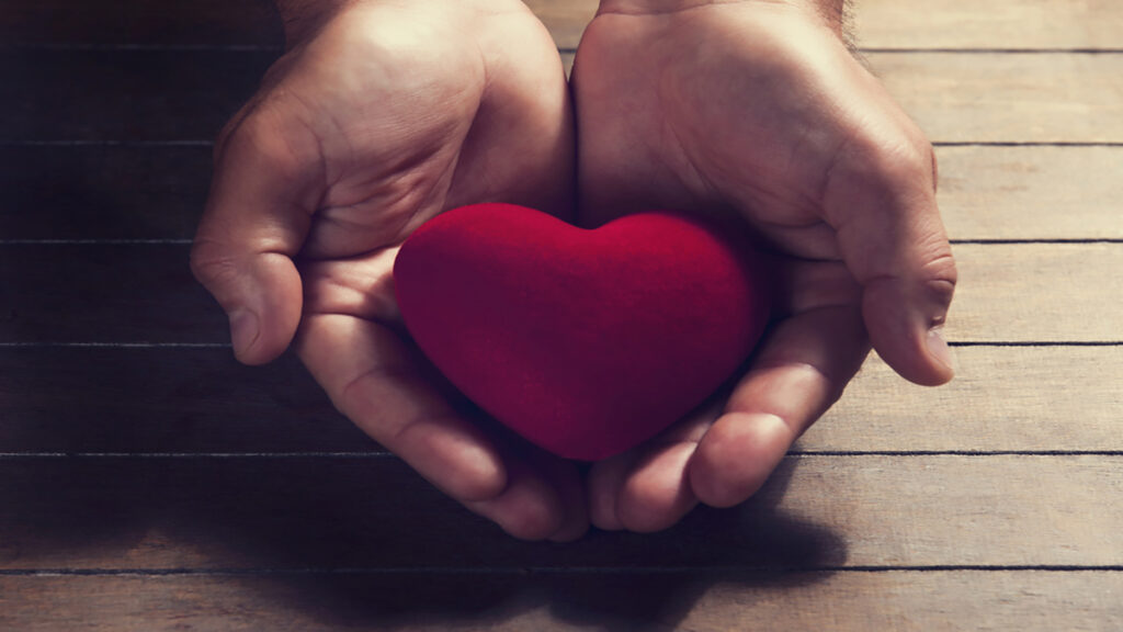 Hands cupping a red heart on a wooden surface, conveying care and compassion.