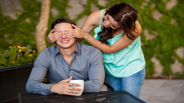 Woman covers a man's eyes from behind as he smiles and holds a mug at an outdoor table in a garden setting.