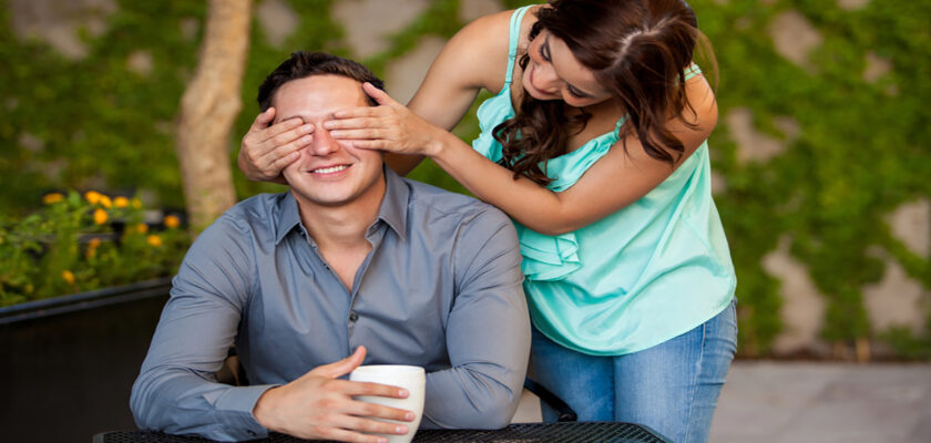 Woman covers a man's eyes from behind as he smiles and holds a mug at an outdoor table in a garden setting.