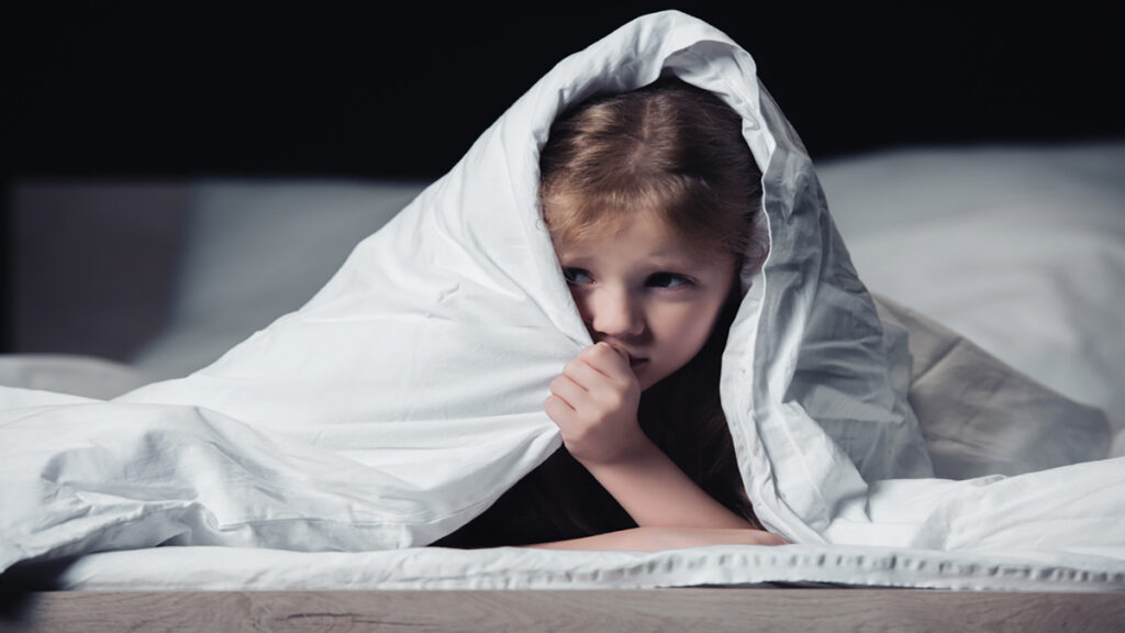 Young girl lying in bed under a white blanket, peeking from beneath it.