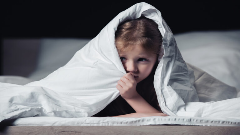 Young girl lying in bed under a white blanket, peeking from beneath it.